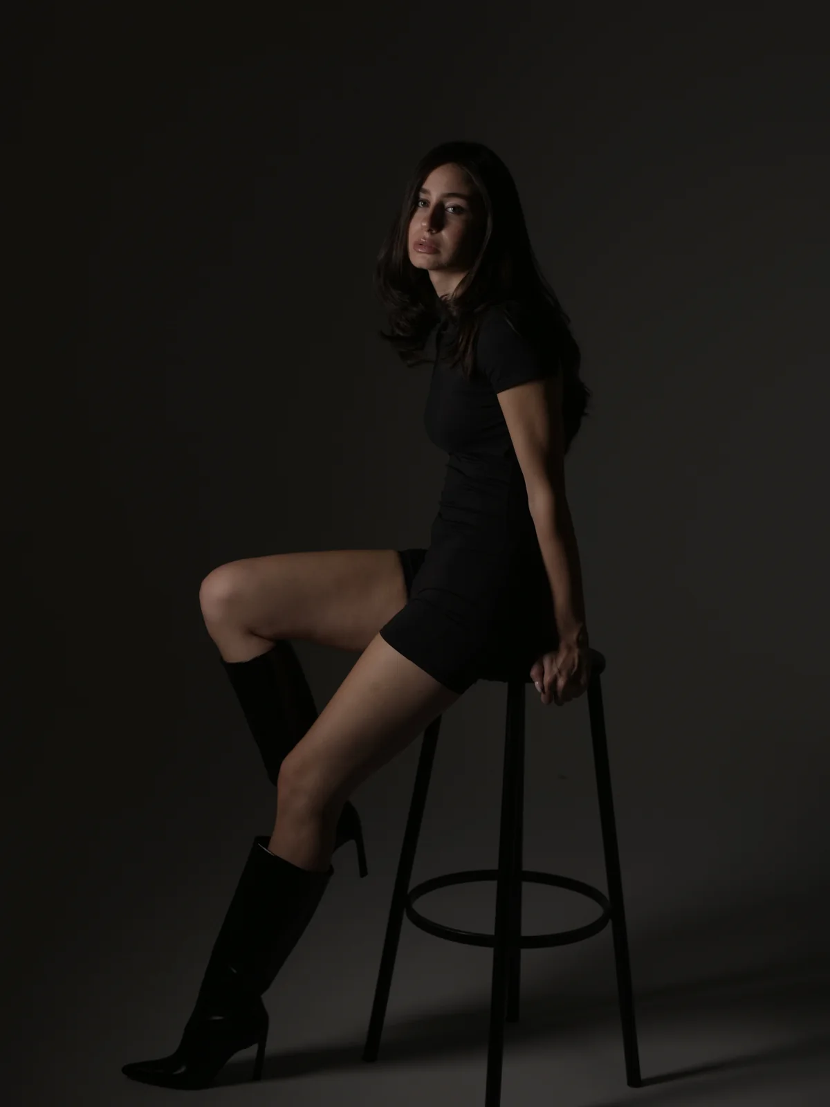 Editorial portrait of Mari seated on a stool in a fitted black dress and boots