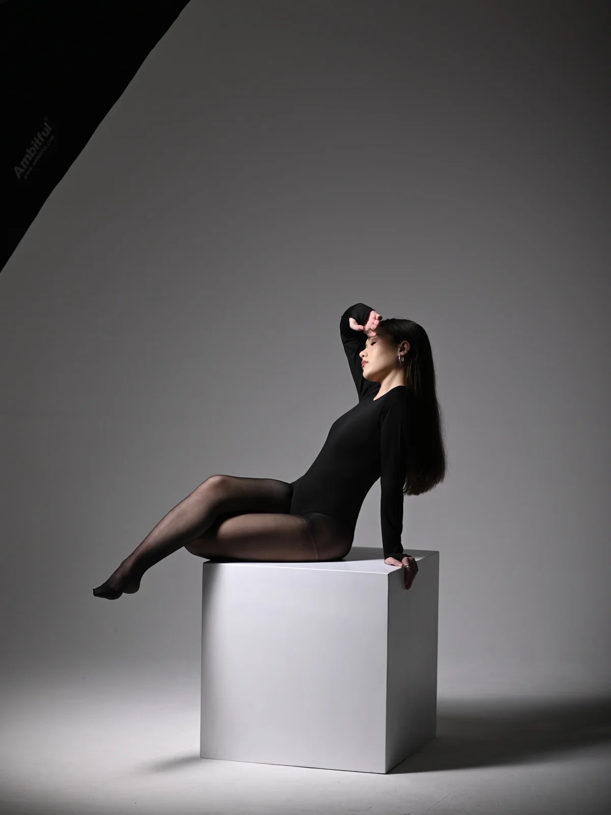Editorial studio portrait of Mariam in a black bodysuit seated on a white cube