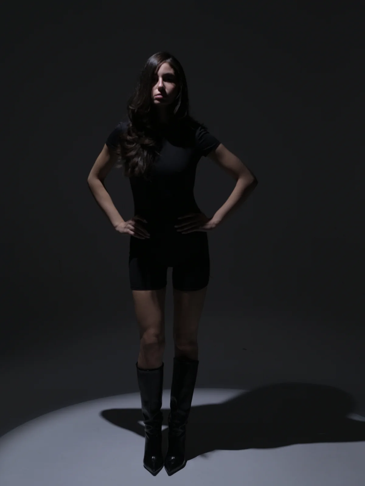 Dramatic spotlight portrait of Mari standing in a black bodysuit and knee-high boots